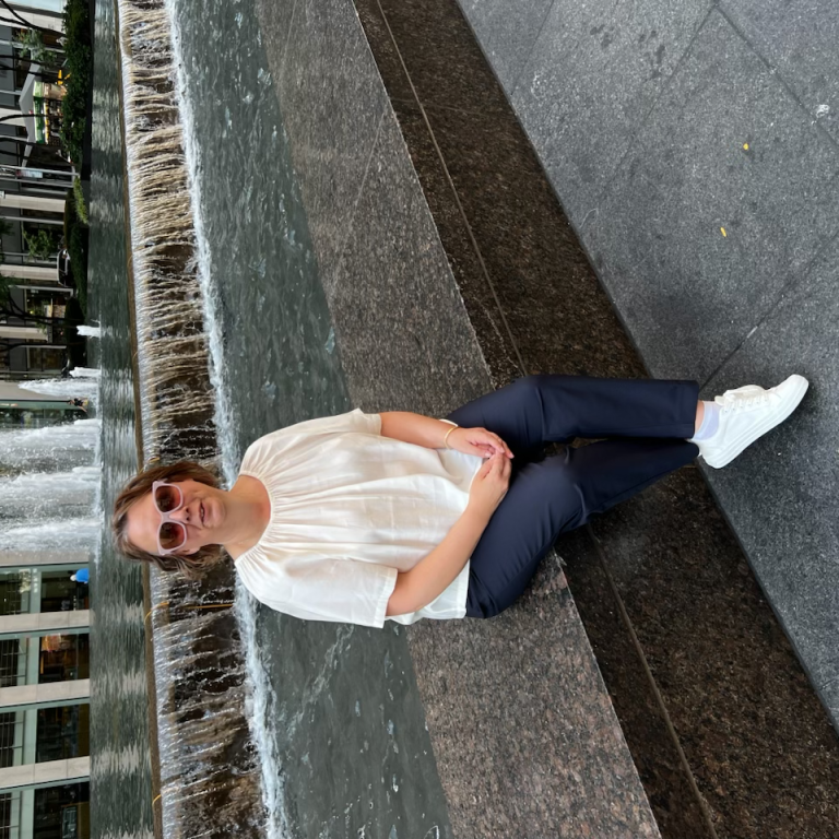 A woman sits by a fountain in an urban setting, wearing sunglasses and a light blouse.