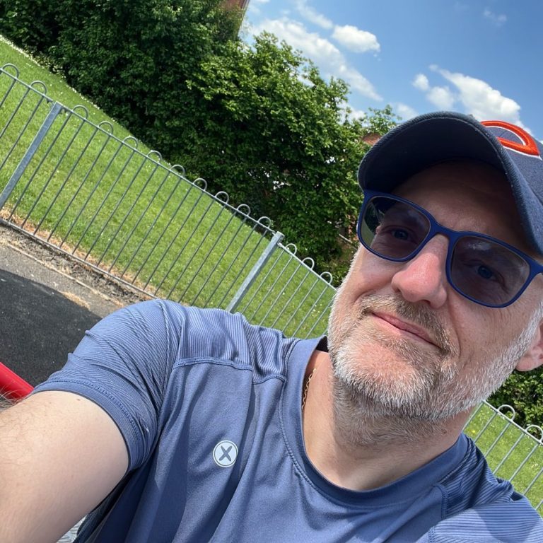Man with glasses and a cap, smiling outdoors in a green park environment.
