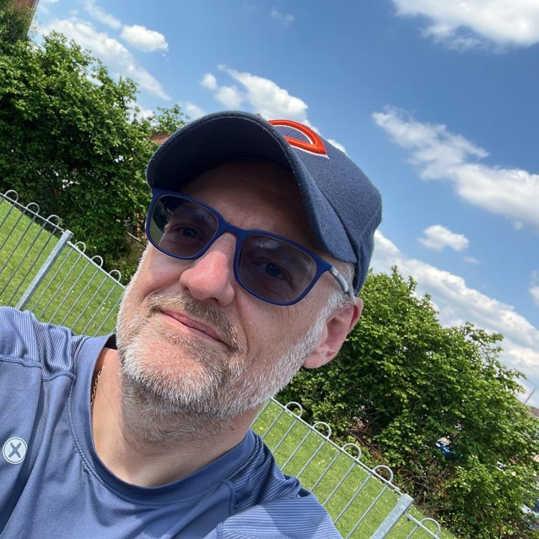 Man with glasses and a cap, smiling outdoors against a sunny sky and greenery.